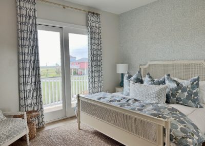 Beach house primary bedroom with patterned curtains, cane bed, layered blue bedding, and balcony doors