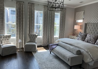 Gray primary bedroom with upholstered bed, lounge chair, patterned drapes, and soft lighting
