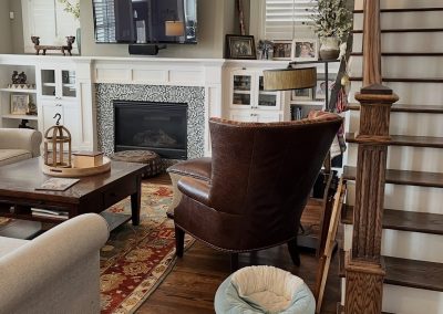 Living room before redesign with fireplace built-ins, dark leather chair, staircase, and patterned rug