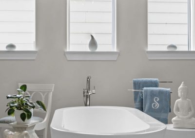 Primary bathroom with freestanding tub, patterned Roman shades, blue monogram towels, and chandelier