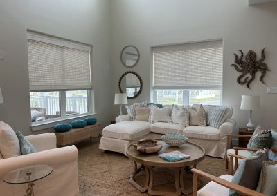 Coastal living room with white sectional, woven bench, blue accents, roller shades, and round coffee table