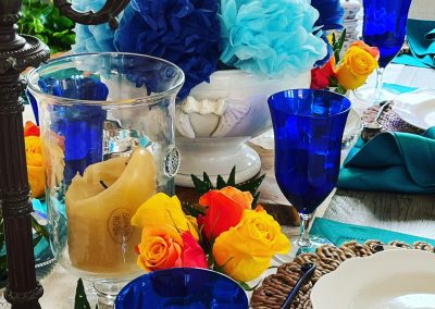 Colorful blue tablescape with woven placemats, cobalt glassware, roses, candles, and shell centerpiece