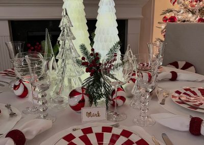 Red and white holiday table setting with peppermint-striped dinnerware, illuminated glass trees, crystal stemware, and Christmas greenery.